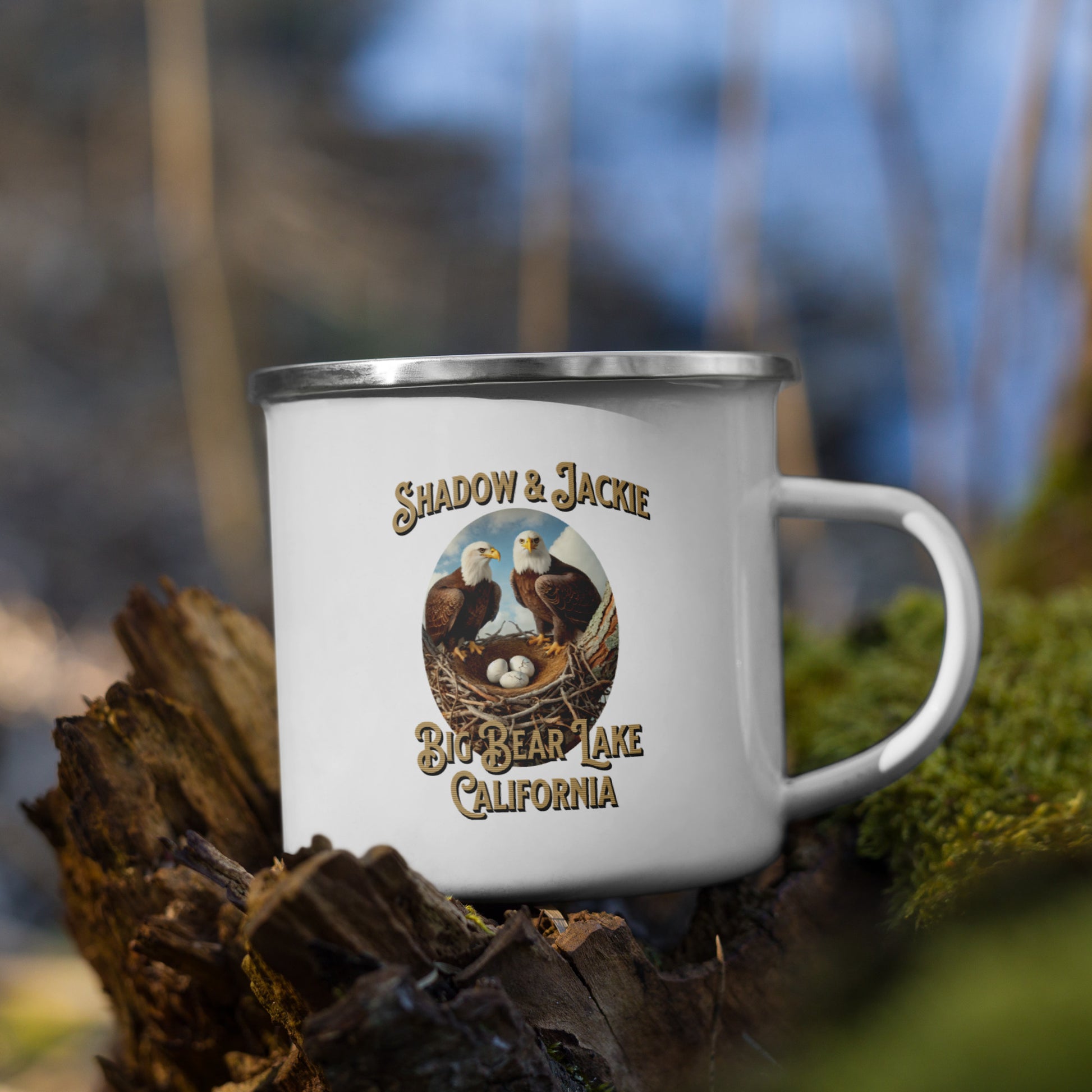 A white enamel mug with silver rim and handle, featuring an image of three baby eagles and text 'Shadow & Jackie Big Bear Lake California', placed on a natural setting with trees in the background.