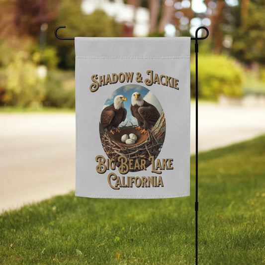 A personalized garden flag featuring an illustration of two bald eagles and three eggs in a nest, with the text 'SHADOW & JACKIE' and 'BIG BEAR LAKE CALIFORNIA'. The flag is displayed on a black metal garden flag stand in a grassy yard with a blurred background.